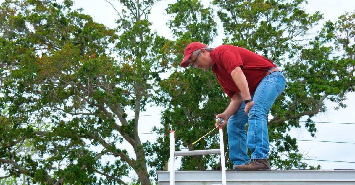 Man in a red shirt stands on a roof beside a ladder, using a tape measure while inspecting the roof edge.