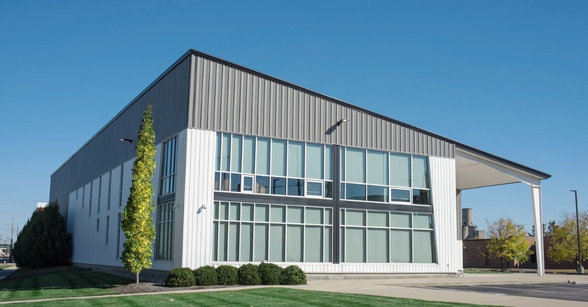 Modern commercial building with a steep sloped metal roof, large windows, white siding, and a green lawn in front.
