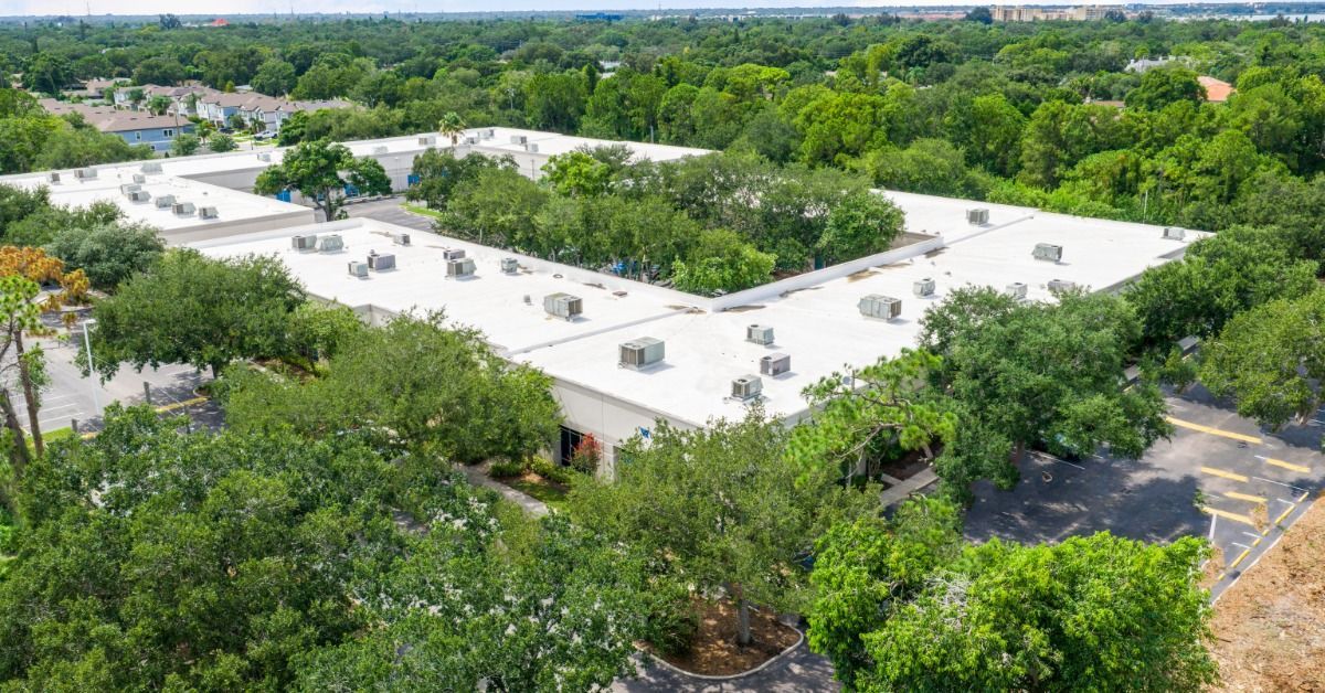 Large white flat roofs with rooftop HVAC units on commercial buildings surrounded by dense green trees and parking lots.