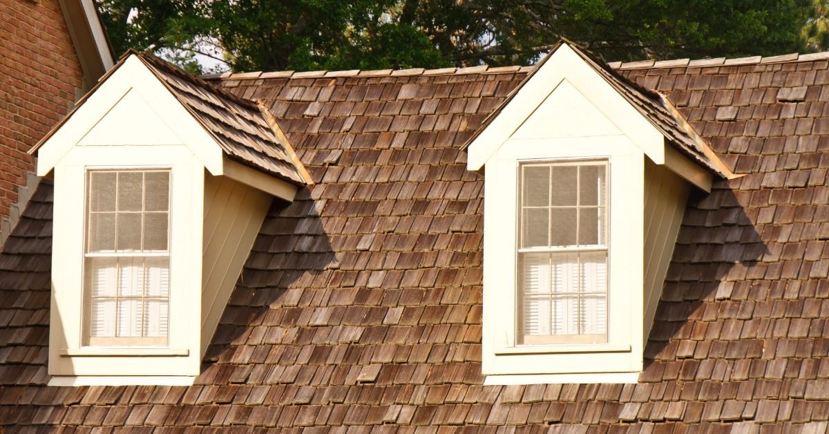 Brown wood shake shingle roof with two white dormer windows, triangular gables, and leafy green trees in the background.