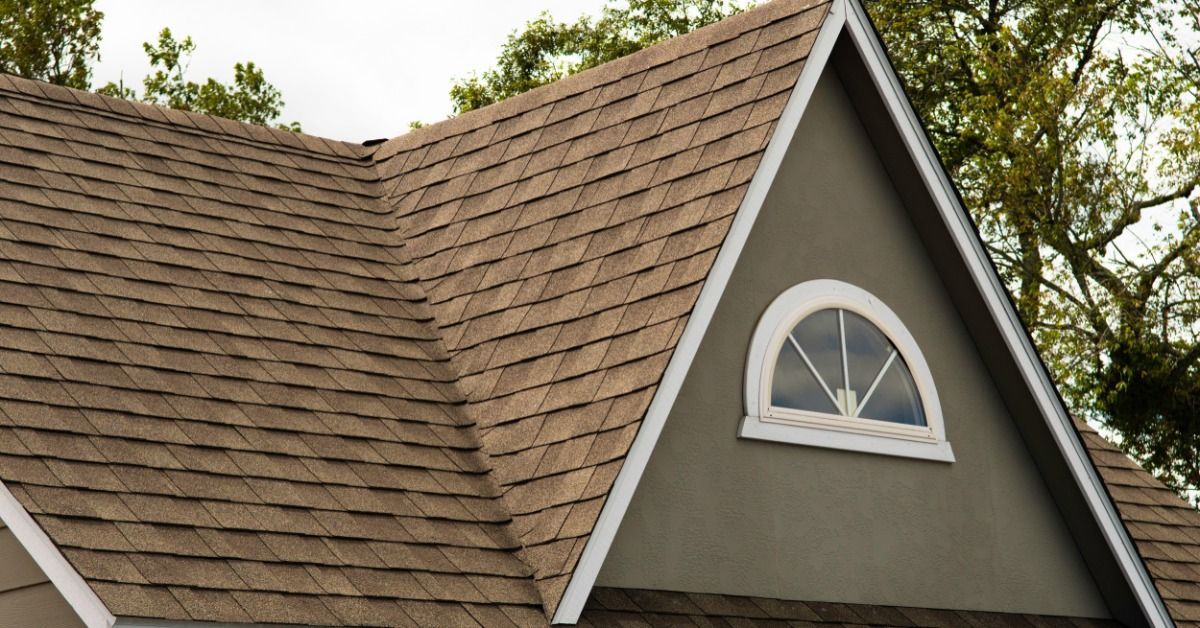 Brown asphalt shingle roof with a steep triangular gray gable, white trim, an arched window, and green trees behind it.
