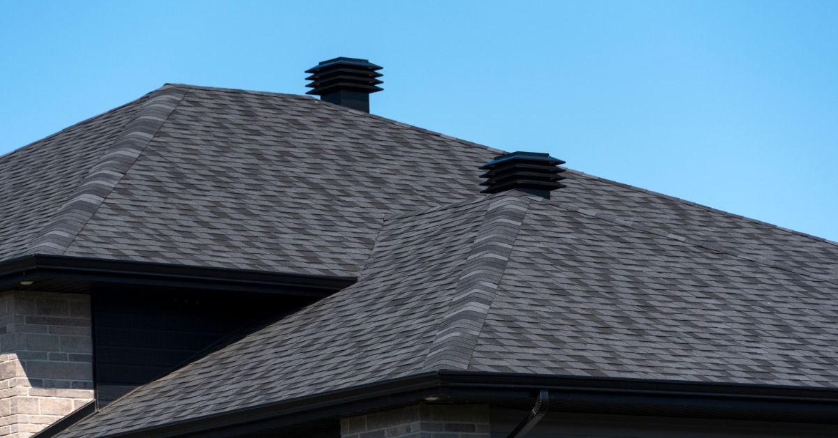 Two black roof vents rise above a gray asphalt shingle roof with multiple slopes, brick walls, and a clear blue sky.
