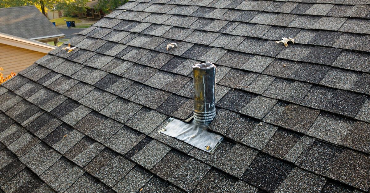A metal vent pipe with flashing protrudes from a dark gray asphalt shingle roof, with a ridge vent and trees behind it.