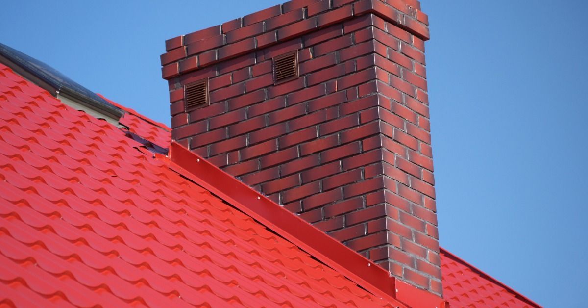 Red brick chimney with two small vents rises from a bright red metal roof, with matching red flashing against a blue sky.