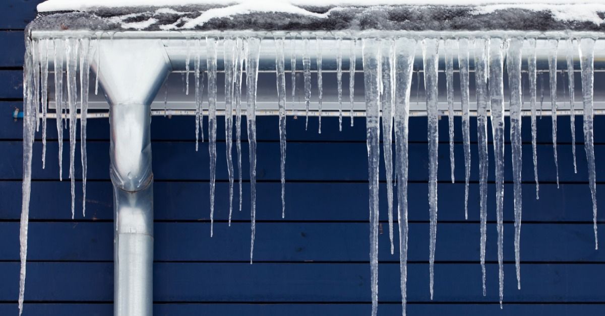 Long icicles hang from a snow-covered metal gutter and downspout along the roofline of a dark blue h