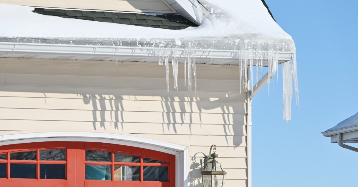 Long icicles hang from a snow-covered garage roof and white gutter above a red garage door on a beige house.