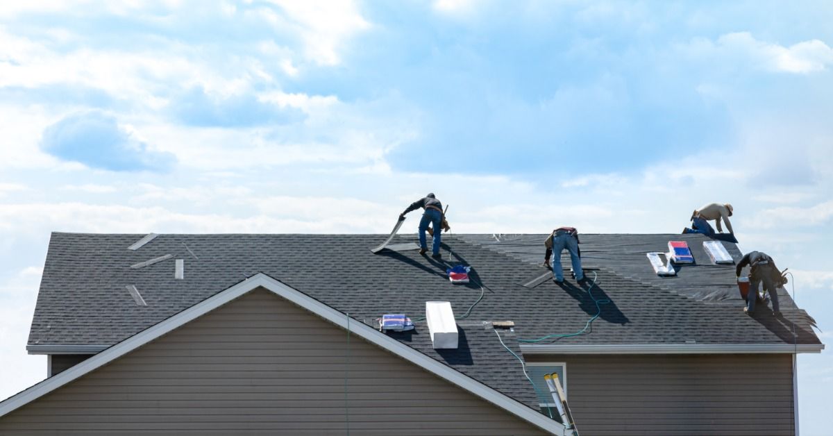 Four roofers install shingles on a residential roof, working with tools and materials under a partly cloudy sky.