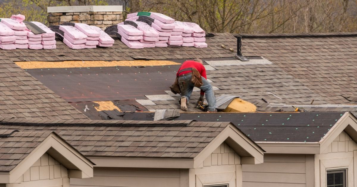 A roofer in a red shirt installs shingles on a residential roof while pink material bundles sit stacked nearby.