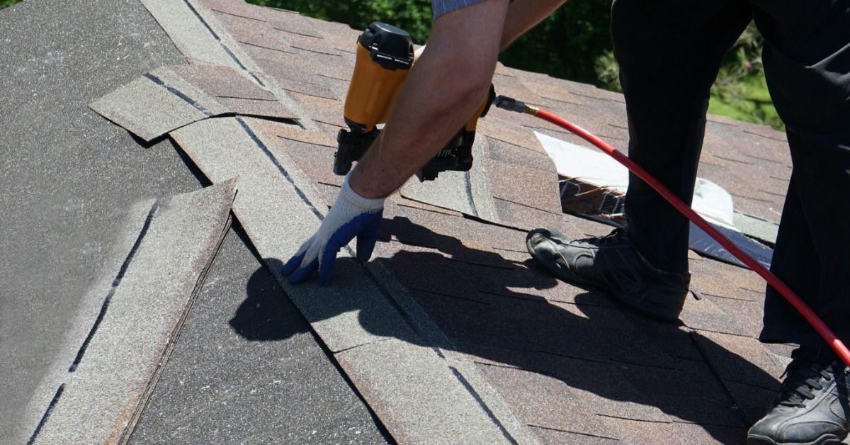 A roofer uses an orange nail gun to install asphalt shingles on a sloped roof during a roofing project.