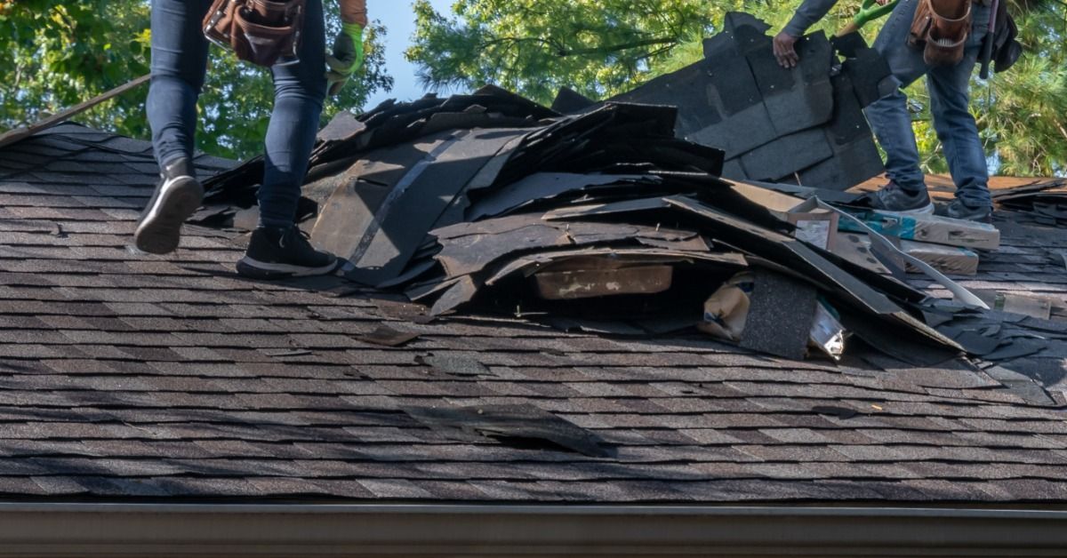 Two roofers remove damaged shingles during a roof tear-off, with debris piled high on a residential rooftop.