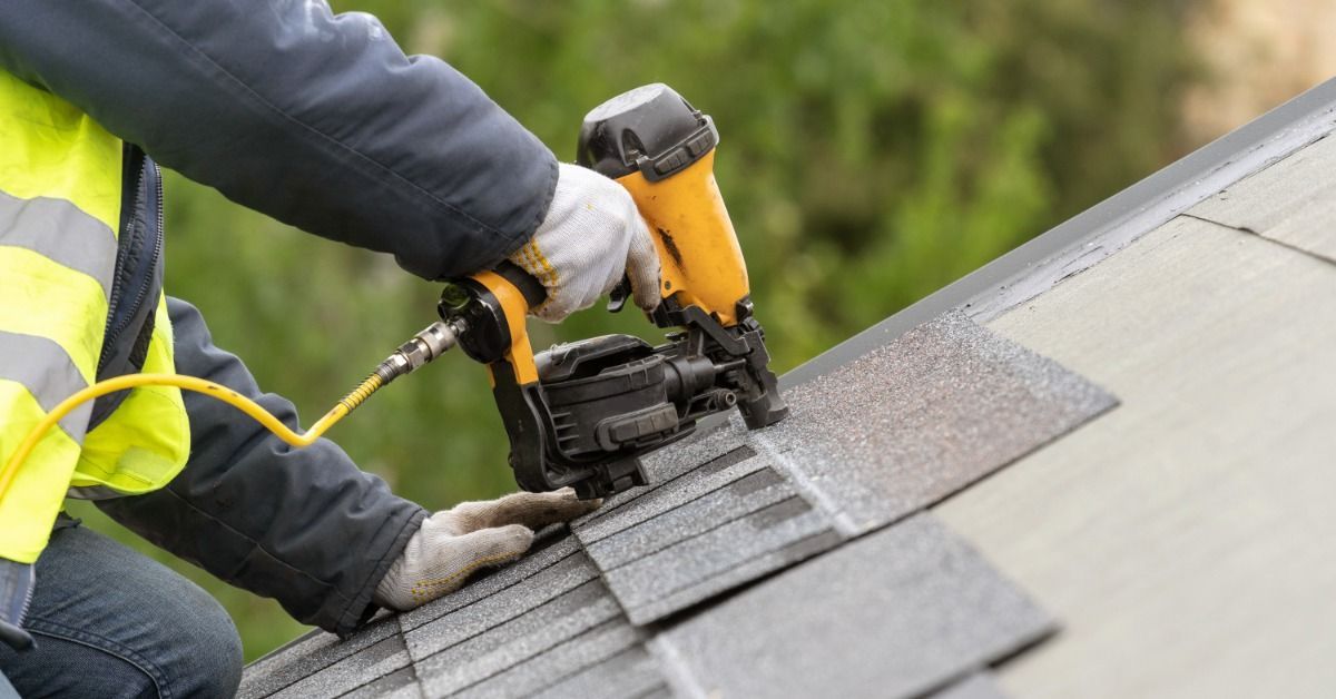 A roofer using a nail gun to install asphalt shingles on a sloped roof, wearing gloves and a safety jacket outdoors.