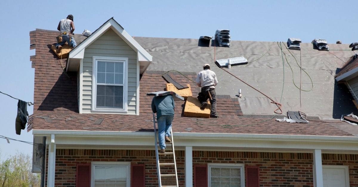 Three roofers replacing shingles on a two-story home, one on a ladder and two on the roof removing old materials.