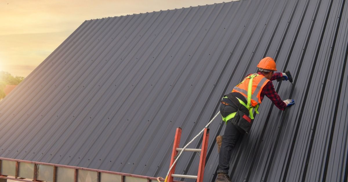 A worker in a safety harness and helmet installs dark metal roofing panels on a steep commercial roof.