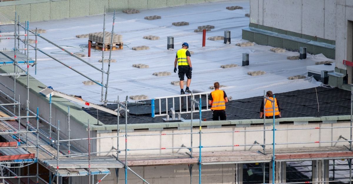 Three workers in safety vests install a flat commercial roofing membrane with scaffolding on a large building.