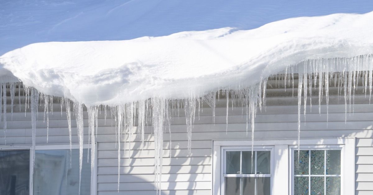 Large icicles hanging from a snow-covered roof edge above house windows during freezing winter weather.