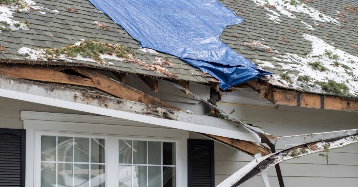 A severely damaged house roof with broken fascia, exposed wood, and a blue tarp covering storm-damaged shingles.
