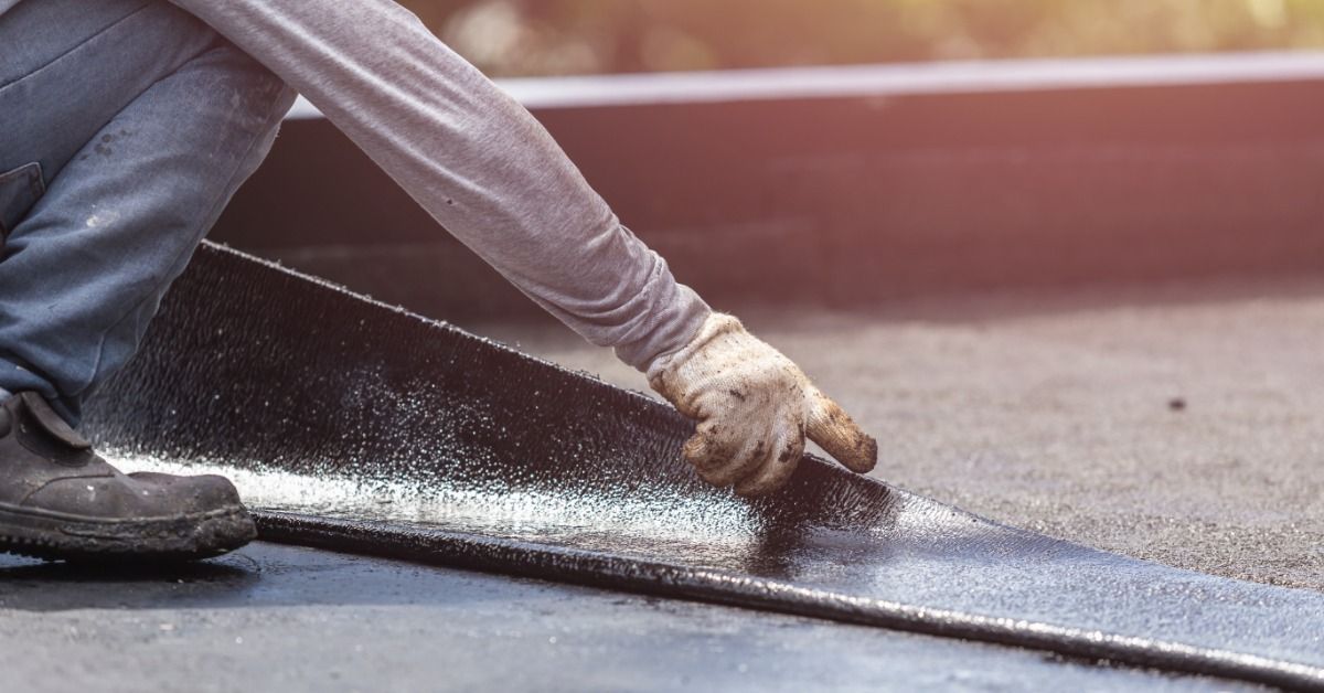 A worker installs a black EPDM roofing membrane across a flat roof surface while securing the material by hand.