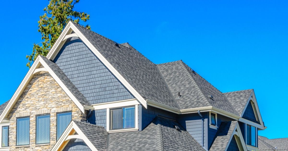 A large gray shingled roof on a two-story home with stone siding beneath a bright blue sky and tall trees.