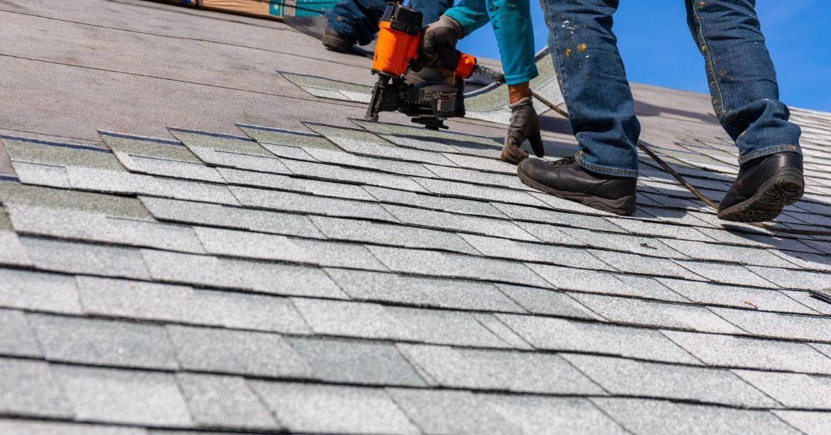 Roofers install gray asphalt shingles on a sloped residential roof using a nail gun under a clear blue sky.