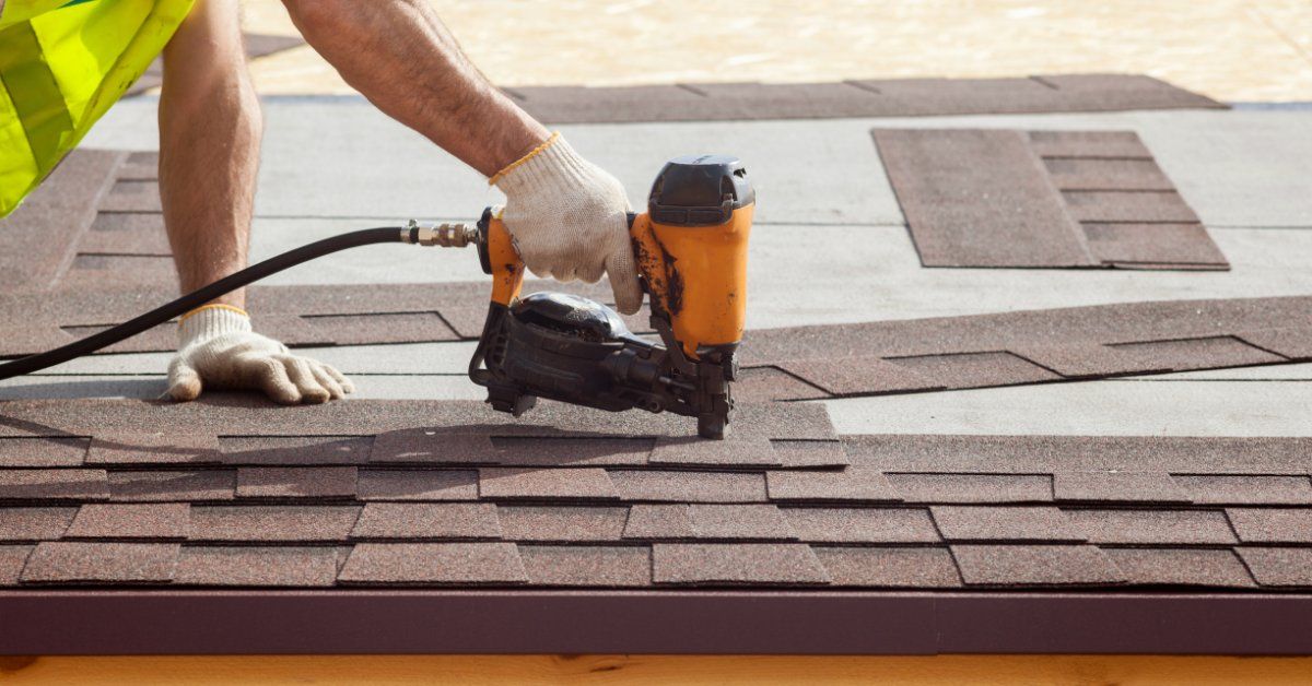 A roofer in a safety vest using a pneumatic nail gun to install brown asphalt shingles on a residential roof deck.