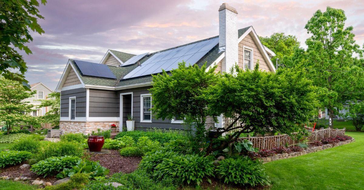 Solar panels line the roof of a gray home surrounded by lush landscaping, mature trees, and a colorful evening sky.