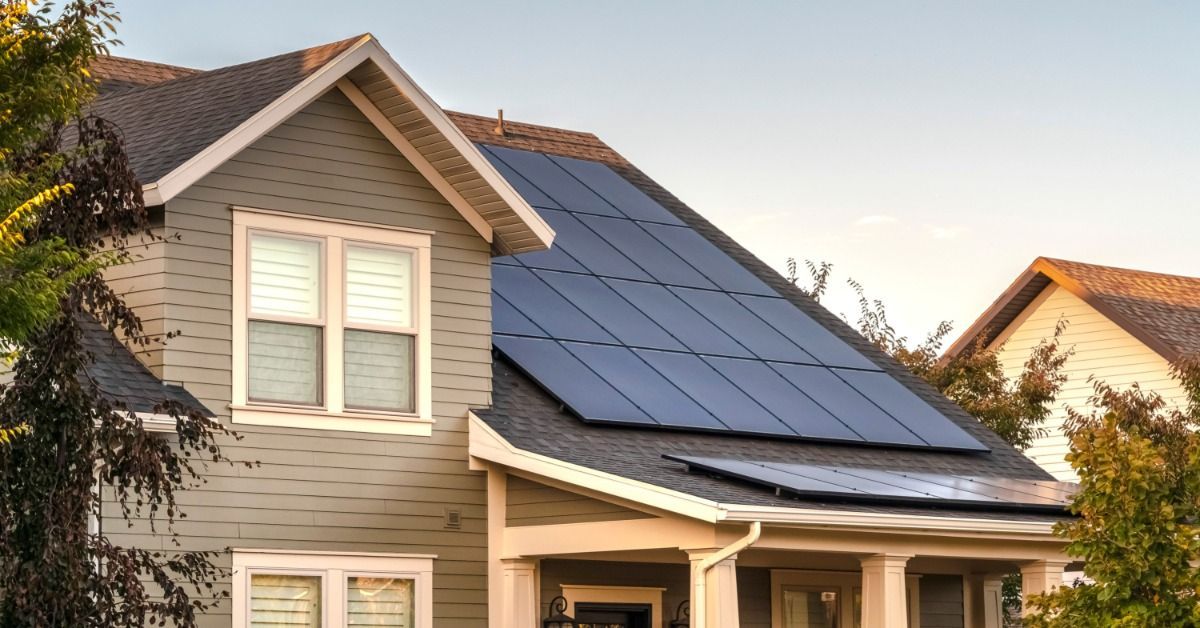 Large solar panels cover the roof of a two-story home with a covered porch, framed by trees in warm evening light.