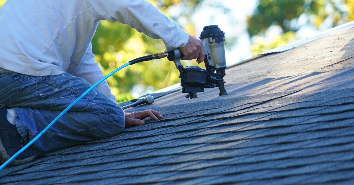 A close-up of a roofer installing asphalt shingles with a pneumatic nail gun on a sloped residential roof.