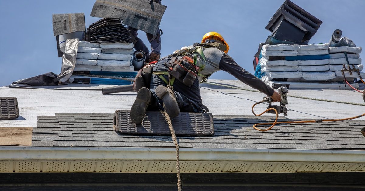Roofers installing asphalt shingles on a residential roof, using safety harnesses and pneumatic nail guns.