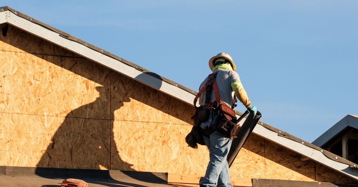 A roofer carrying materials across a roof deck during construction, with plywood walls, safety gear,