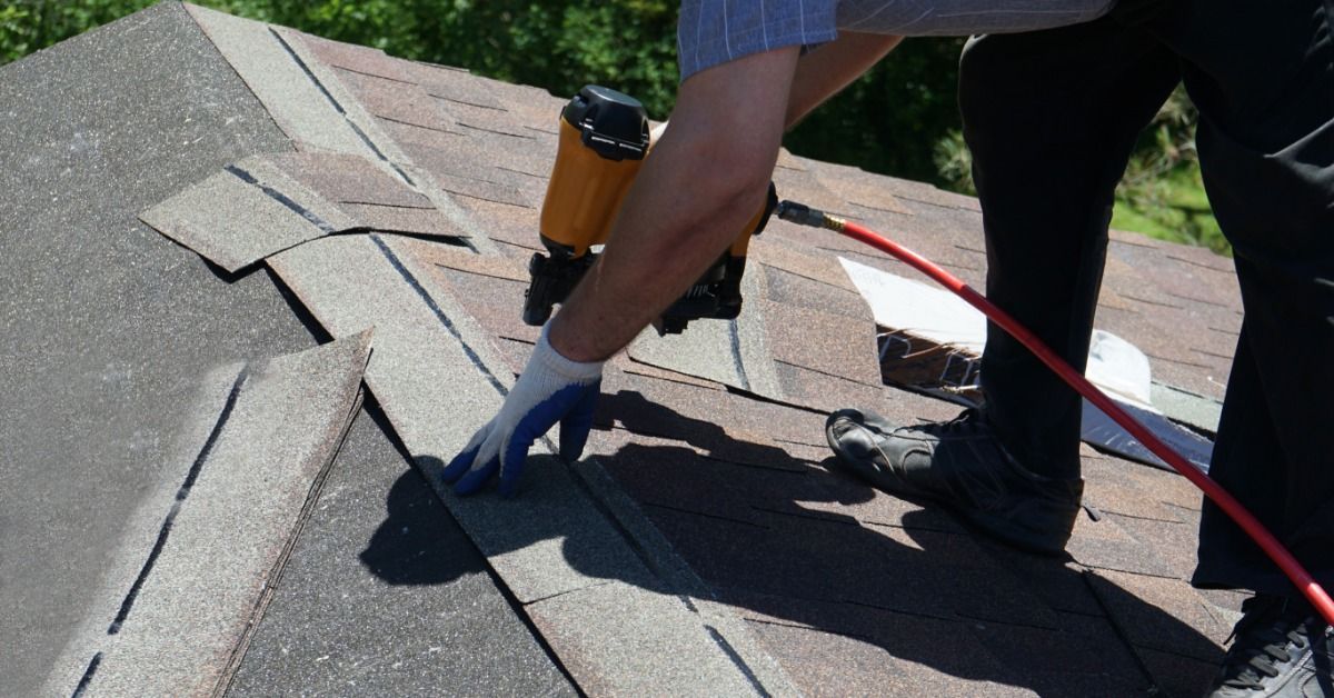 A close-up of a roofer installing asphalt shingles with a nail gun on a sloped roof during replacement work.
