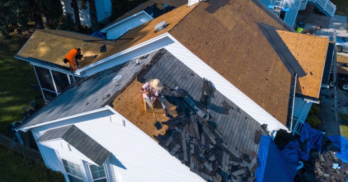 An aerial view of roof replacement on a white house, with workers removing shingles and exposed deck