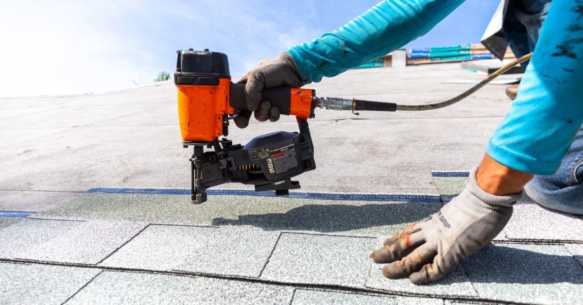 A roofing contractor uses a pneumatic nail gun to install gray asphalt shingles on a residential roof under clear skies.