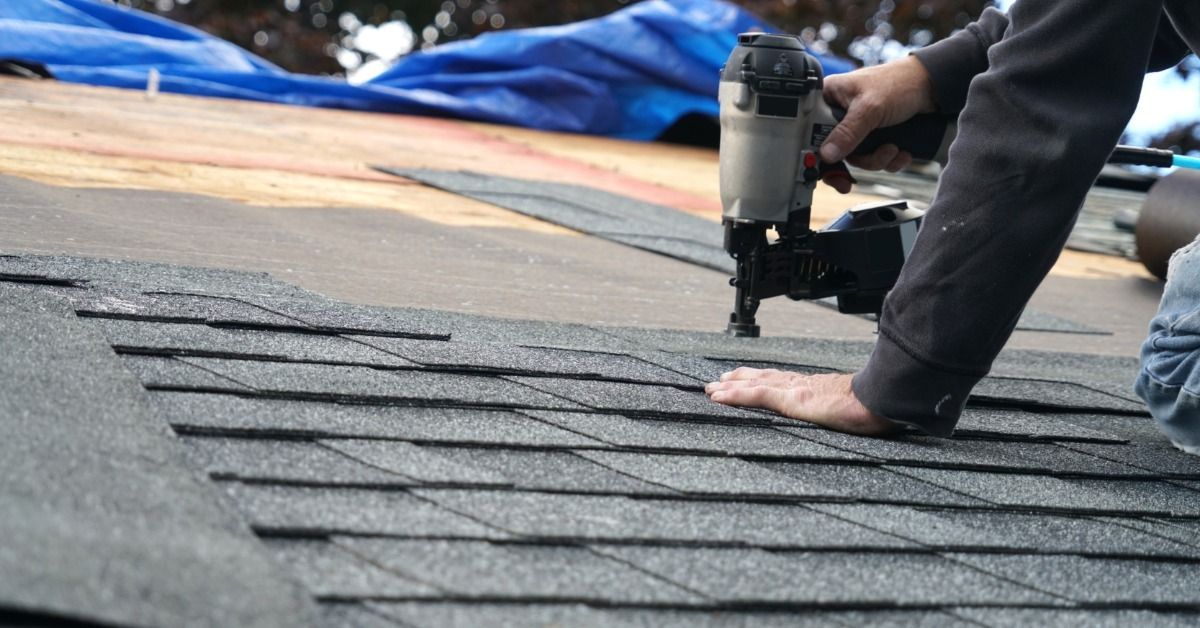 A close-up of a roofer installing asphalt shingles with a pneumatic nail gun on a roof deck during a roofing project.