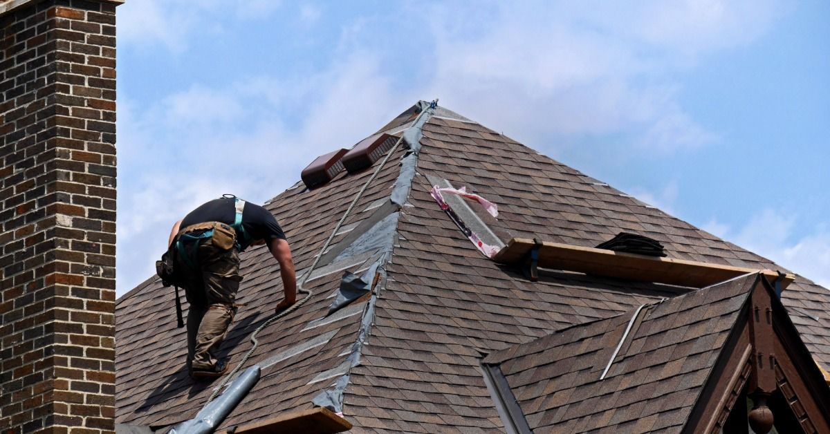 A roofer repairs a steep shingle roof beside a brick chimney, replacing flashing beneath a bright blue sky.