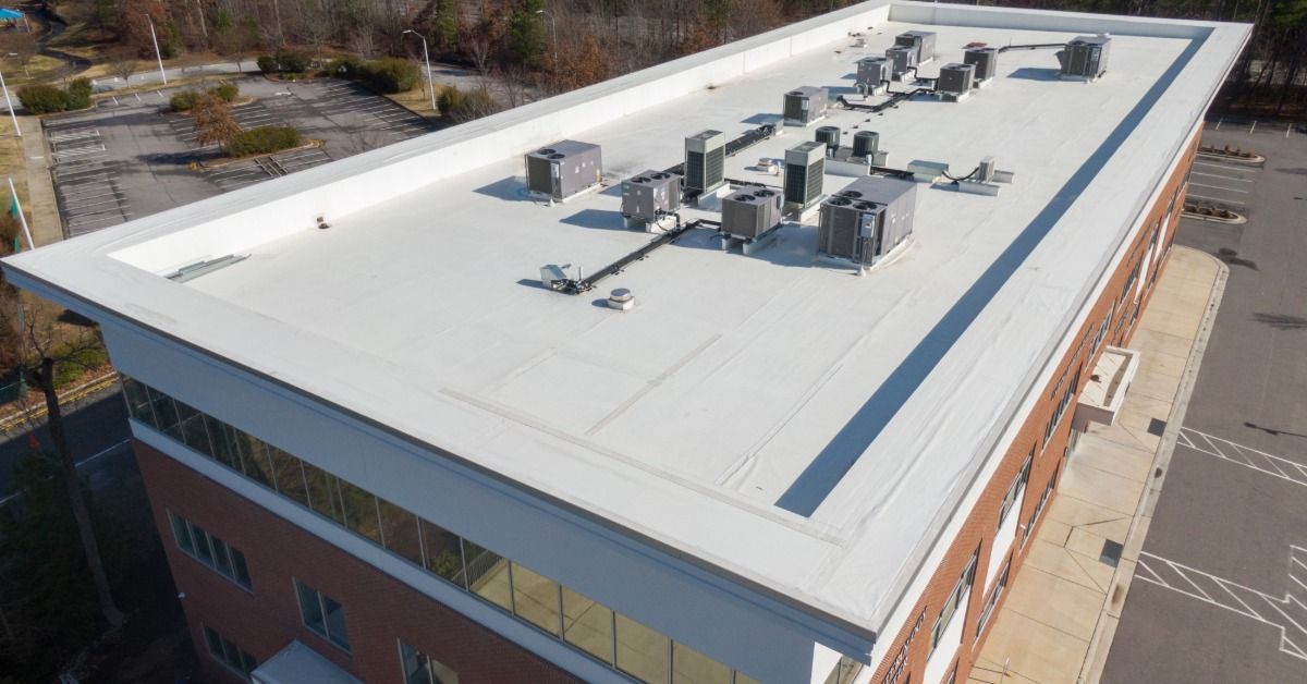 An aerial view of a flat white commercial roof with HVAC units atop a brick office building beside nearby parking lots.