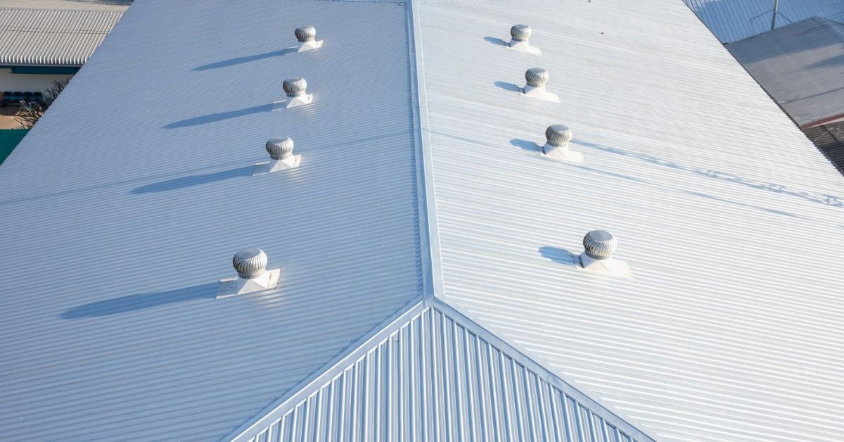 An aerial view of a white corrugated metal commercial roof with multiple turbine vents casting long