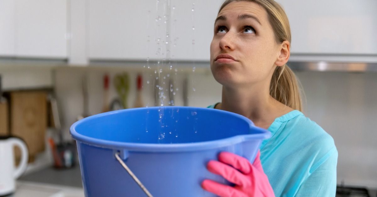 Woman holding a blue bucket beneath a ceiling leak in her kitchen, catching dripping water while looking up with concern.