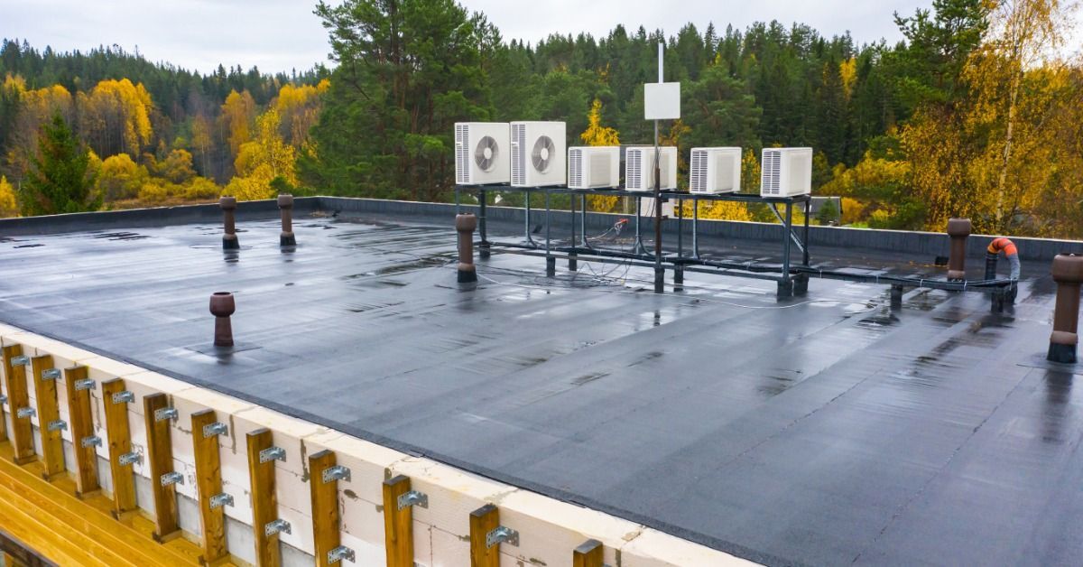 Flat commercial roof with black membrane, rooftop HVAC units, vent pipes, and a forested autumn land