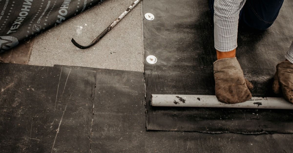 Worker installing black flat roofing membrane around a roof pipe, sealing seams for a durable waterproof surface.