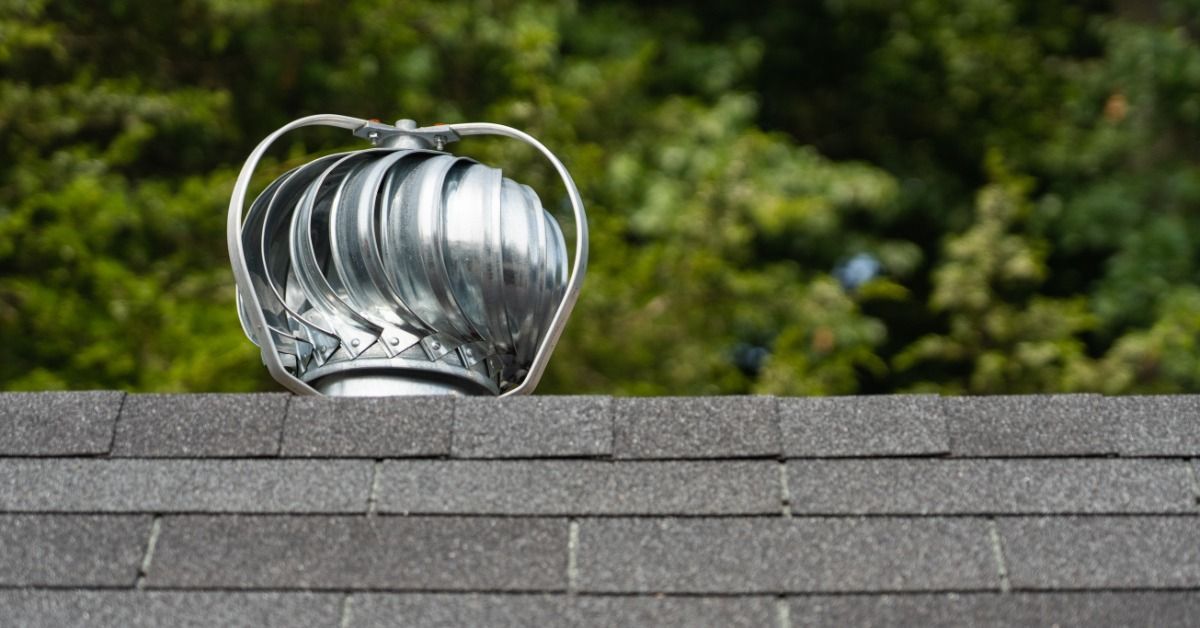Silver rooftop turbine vent mounted on a gray shingle roof with green trees in the background on a bright sunny day.