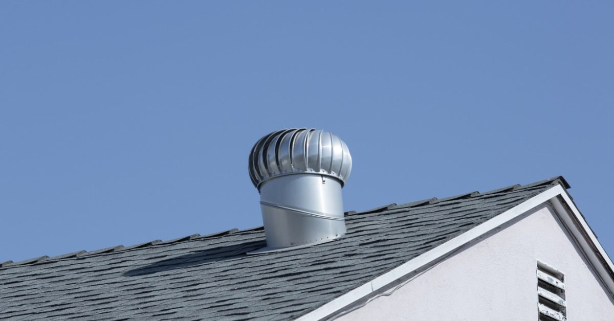 Silver rooftop turbine vent mounted on a gray shingle roof beneath a clear blue sky above the white