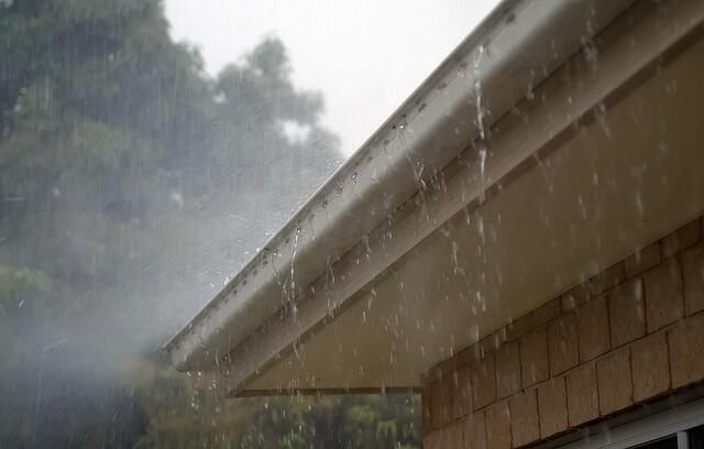 Rain falling from a roof's gutter, gray sky, beige trim, wooden siding.