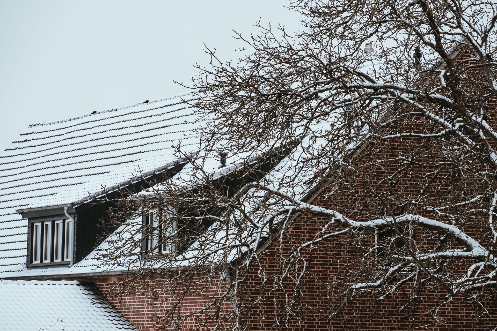 Snow-covered brick house with a dormer and bare tree branches against a cloudy sky.