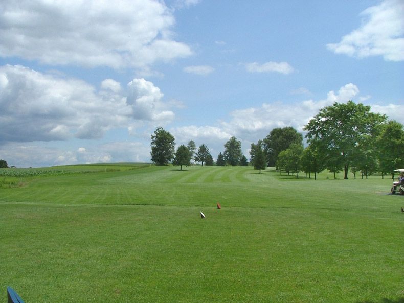 Wide green golf course under a partly cloudy blue sky, with trees lining the fairway.