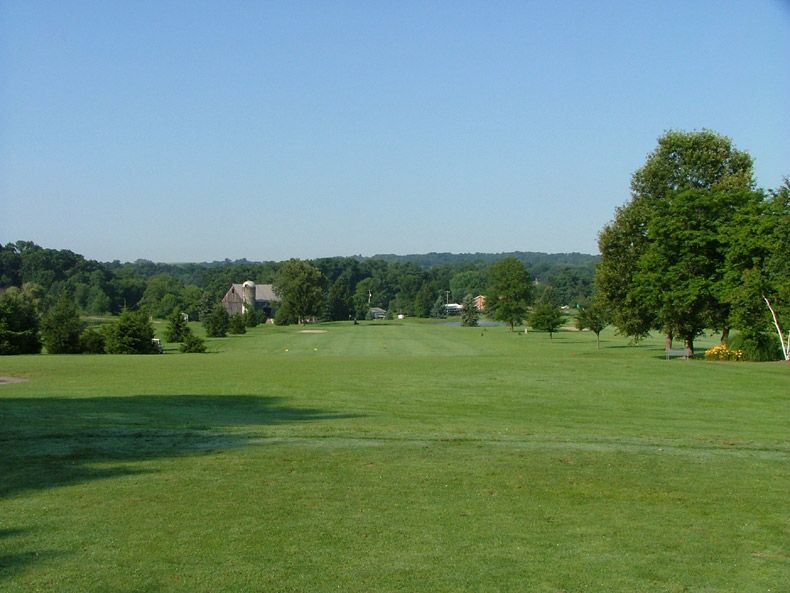 Wide green grassy field under a blue sky with trees and distant houses in the background