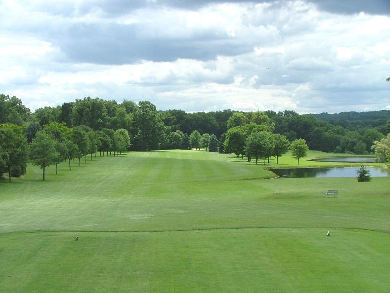 Golf course fairway under cloudy sky, with trees lining the green and a small pond on the right.