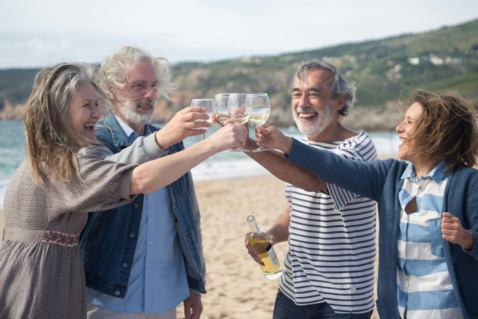 Four people toasting drinks on a beach, smiling. Blue water and sky in background.