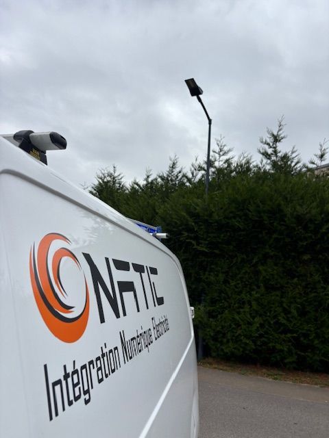 Camionnette blanche avec le logo « ONATIC », lampadaire et haie verte sous un ciel nuageux.