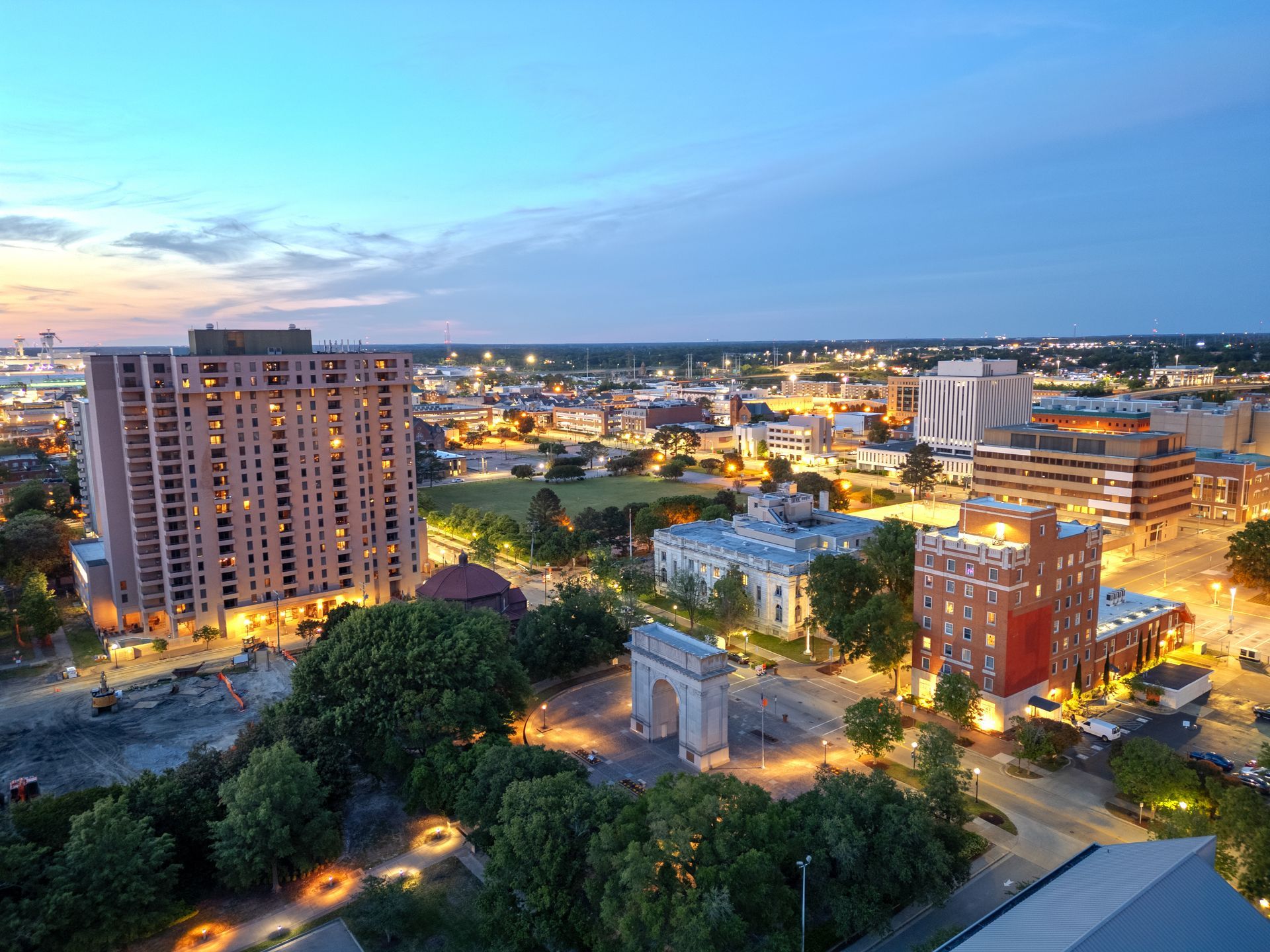 City skyline at dusk with lit buildings, trees, and a blue-orange sunset sky