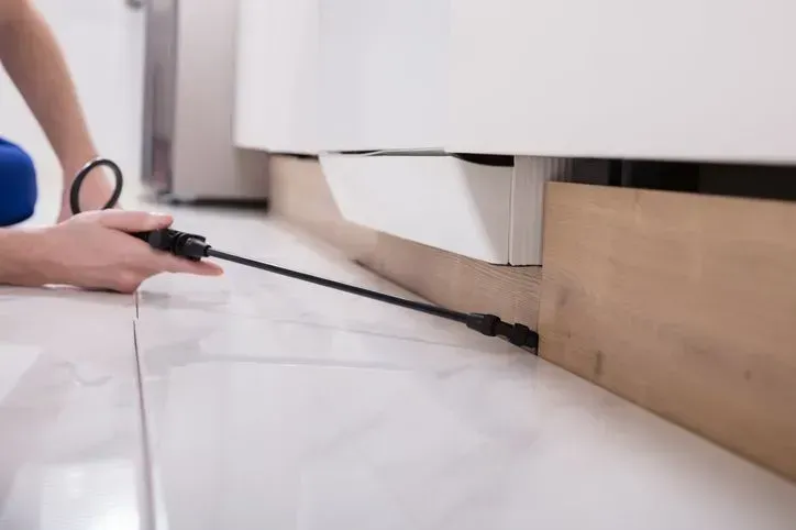 Person cleaning under a cabinet with a vacuum nozzle on a white floor